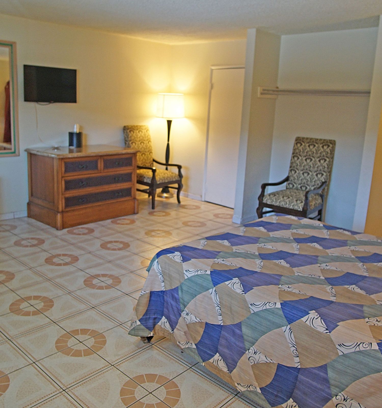 A hotel room with a bed, patterned bedspread, armchair, lamp, TV, dresser, mirror, mini fridge, and an open bathroom with a tiled shower.