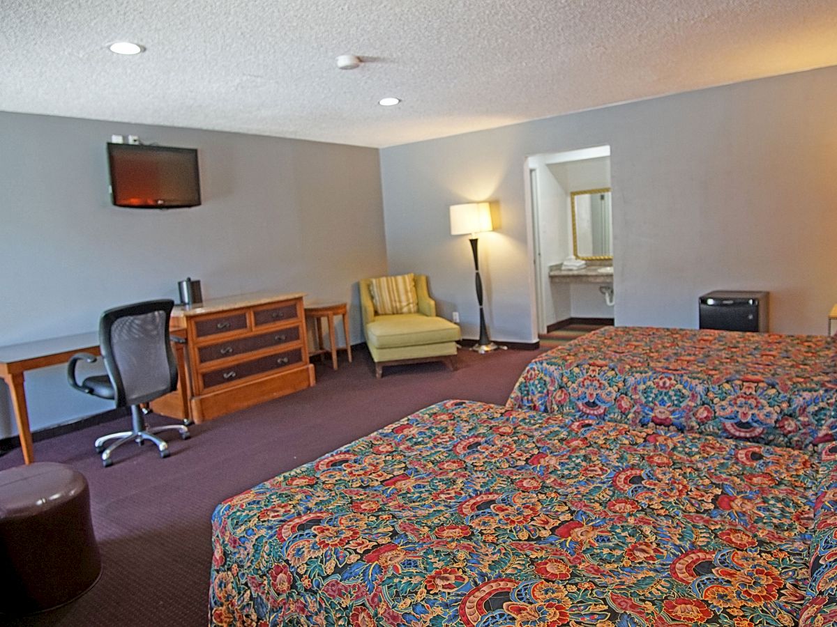 A hotel room with two beds, desk, chair, TV, two lamps, armchair, and a mini-fridge. There is a sink area visible in the background.