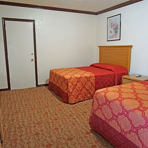 The image shows a hotel room with two beds, a wall-mounted TV, a small desk, two chairs, and a picture on the wall.