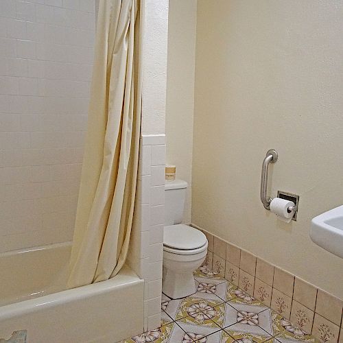 The image depicts a small bathroom with a bathtub, shower curtain, toilet, wall-mounted sink, mirror, and tiled floor.