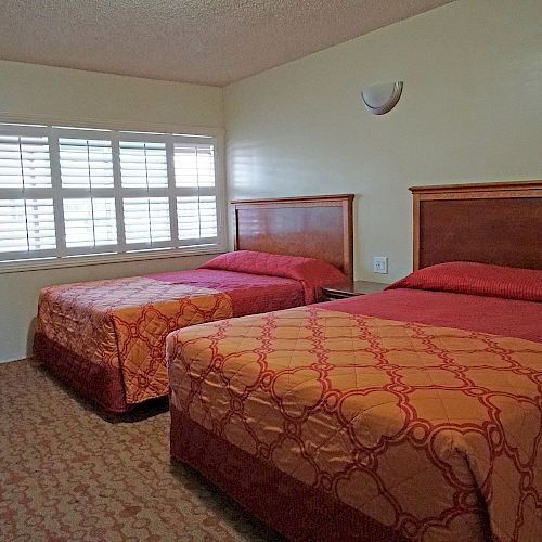 The image shows a room with two double beds, each with red and orange bedding, a beige carpet, a pink door, and a window with white shutters.