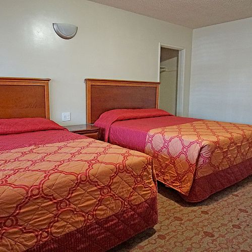 The image shows a hotel room with two double beds covered in patterned bedspreads; a doorway in the back leads to a bathroom.