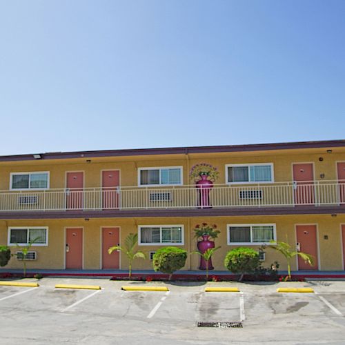 An image of a beige two-story motel with red doors, potted plants, and an empty parking lot in front under a clear blue sky.