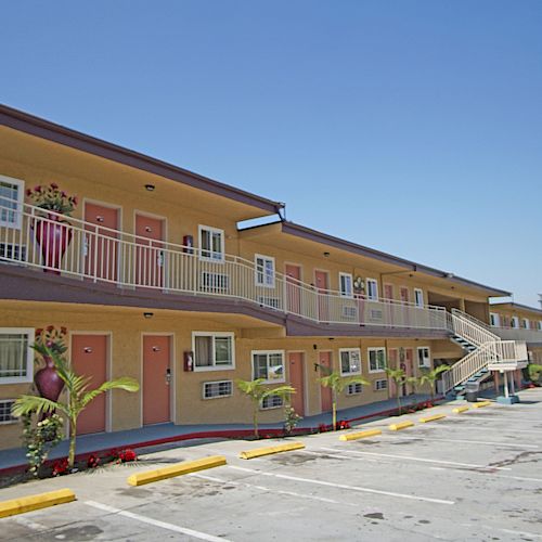 The image shows an exterior view of a two-story motel with beige walls, red doors, a parking lot, and some small plants near the building.