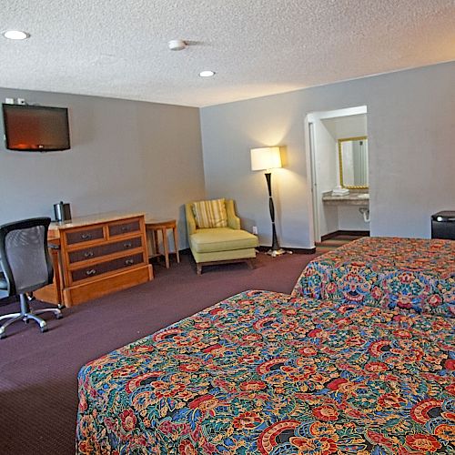 The image shows a double-bed hotel room with a TV, desk, chair, armchair, lamp, and dresser. The beds have colorful bedspreads, and there's a bathroom.