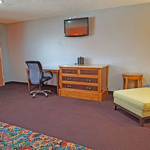 The image shows a hotel room with a desk, office chair, wall-mounted TV, lounge chair, side tables, and a door. The carpet and bed coverings are patterned.