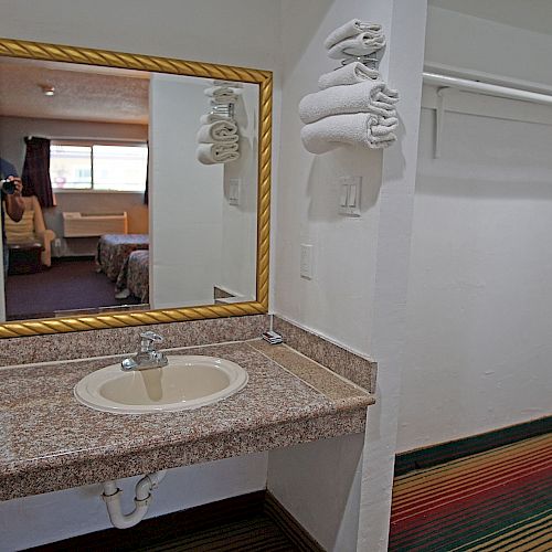 The image shows a hotel room with a sink and mirror area, towel rack, closet, and a view into a bedroom with a seated person.