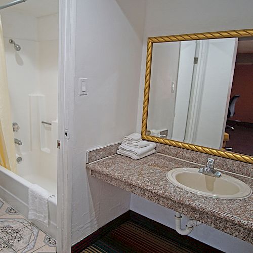 A bathroom with a sink, large mirror, towels, toilet, and shower with a curtain, adjacent to a room with a chair in the background.