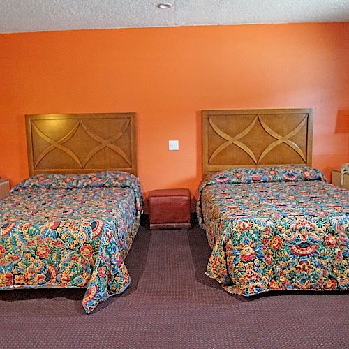 The image shows a hotel room with two double beds, colorful bedspreads, nightstands with lamps, and an orange accent wall in the background.