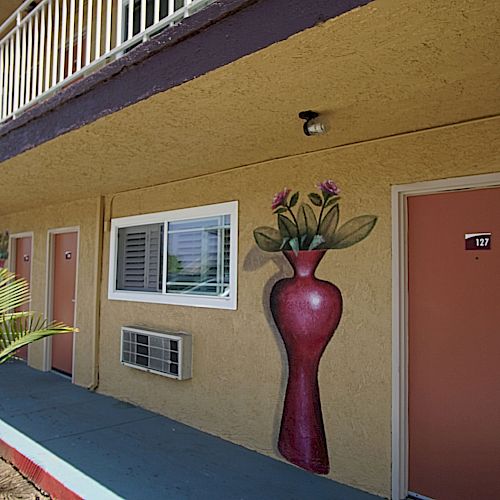 A motel exterior with beige walls, rooms with pink doors, an air conditioning unit, a painted vase with flowers, and a palm plant along the walkway.