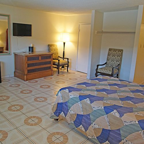 This image shows a hotel room with a bed, patterned bedspread, two chairs, a lamp, dresser, TV, mini-fridge, and an open bathroom area at the back.