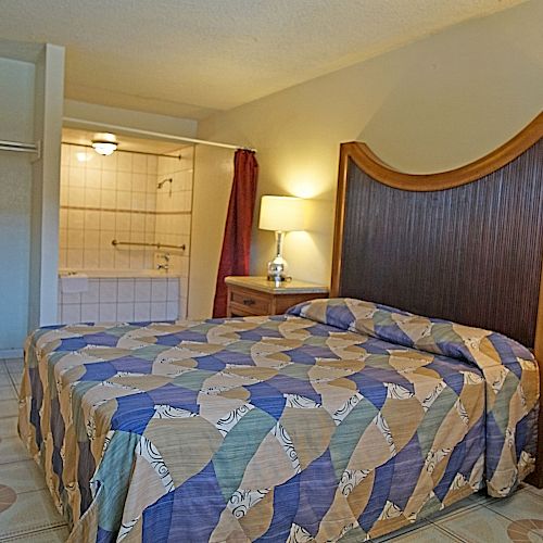 This image shows a cozy hotel room featuring a bed with a colorful geometric bedspread, a chair, a nightstand with a lamp, and an open bathroom.