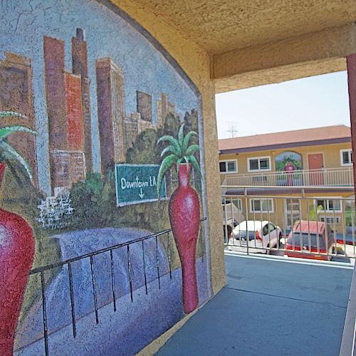 The image features a mural with a cityscape and two red vases, located on an exterior wall of a motel with visible rooms and parked cars in the background.