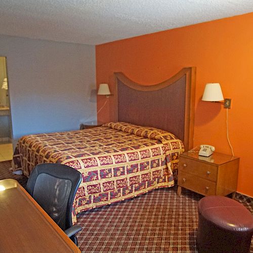 A hotel room with an orange accent wall, a bed, nightstands, a desk with a chair, an armchair, and a visible bathroom area.