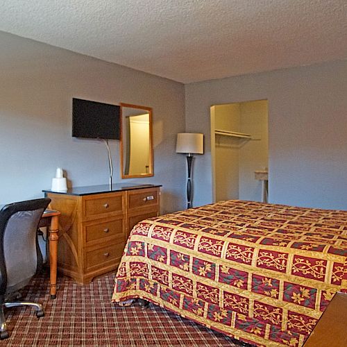 The image shows a modest hotel room with a bed, a desk, a chair, a TV on the wall, and a dresser. The room has a simple design.