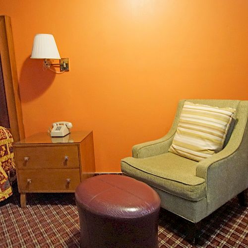 The image shows a hotel room with an orange wall, a bed, a nightstand with a phone, a green chair with a pillow, and an air conditioner.