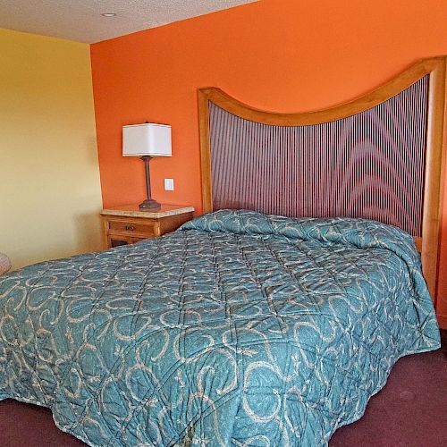 The image shows a bedroom with an orange and yellow wall, a double bed with a patterned blue blanket, two bedside tables with lamps, and an armchair.