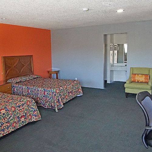 The image shows a hotel room with two beds, a desk, a chair, a green armchair, and a lamp, with an orange accent wall and a view into a bathroom area.