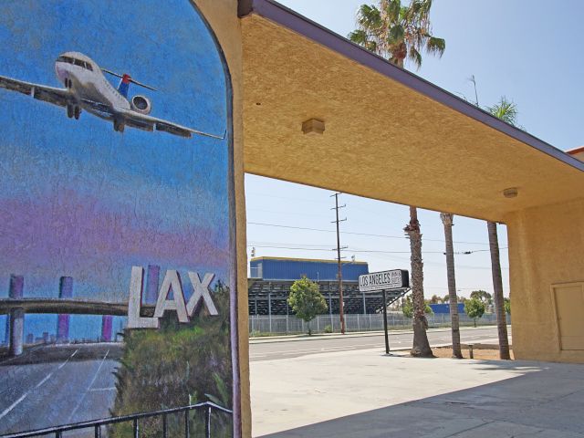 A mural of an airplane flying over a road with the letters LAX on a building next to a covered area with palm trees.