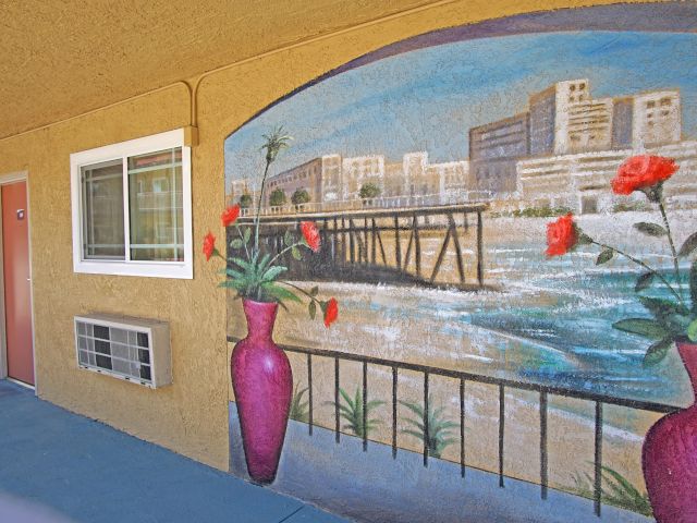 A mural of a beach scene with a pier and city skyline is painted on the exterior wall of a building, accompanied by a window, door, and two vases with flowers.