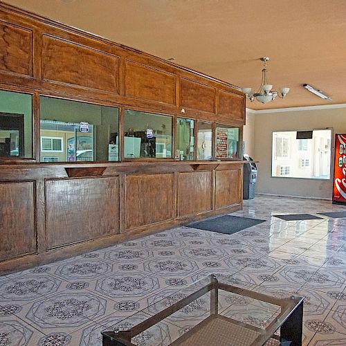 The image shows a hotel reception area with a wooden counter, tiled flooring, chairs, a vending machine, and a door leading outside.