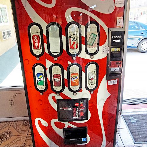 This image shows a red Coca-Cola vending machine with various soft drink options in bottles, located indoors. Thank you.