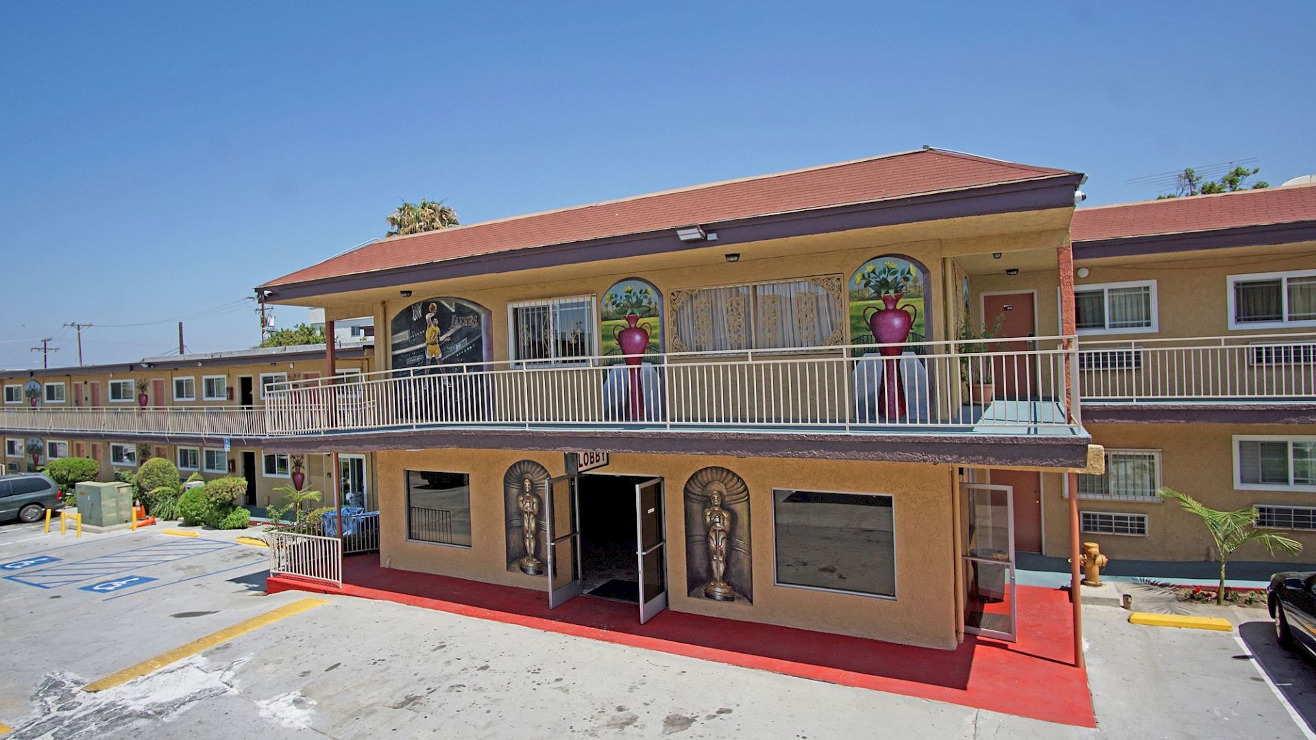 An image of a two-story motel with decorative statues, plants in large pots, red walkway, and parking spaces in front of the building under a clear sky.