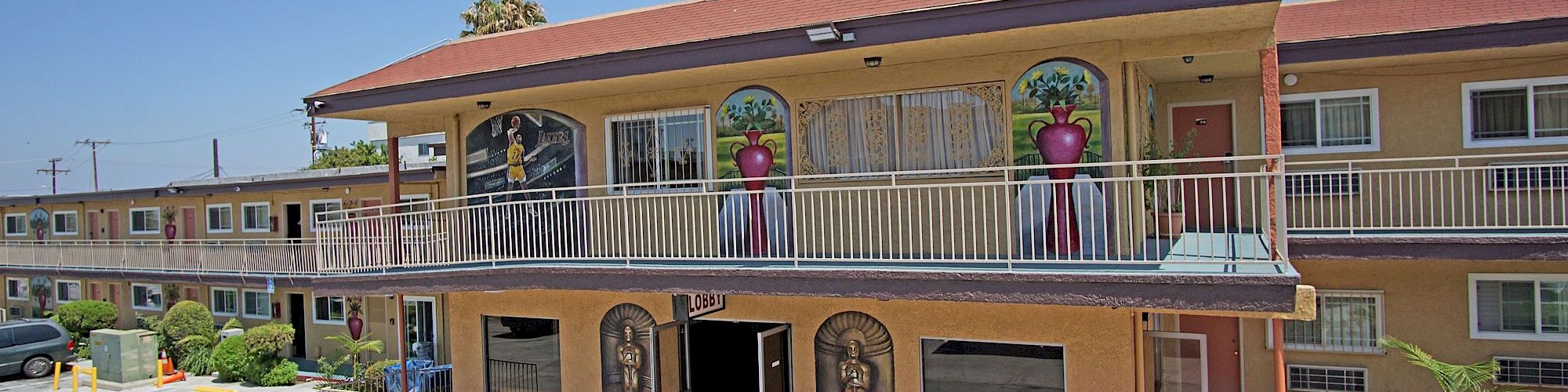 An image of a two-story motel with decorative statues, plants in large pots, red walkway, and parking spaces in front of the building under a clear sky.