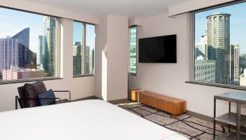 A modern hotel room with large windows featuring city views, a wall-mounted TV, a chair, and light-colored decor.
