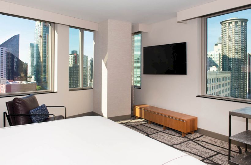 A modern hotel room with large windows featuring city views, a wall-mounted TV, a chair, and light-colored decor.