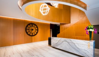 A modern, stylish reception area with a marble countertop, wooden spiral structure, large wall clock, and a vase with colorful flowers.