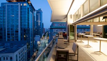 A modern balcony at dusk with tables and chairs, overlooking a cityscape with tall buildings and a busy street below, illuminated by streetlights.