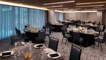 The image shows a dimly-lit banquet hall with multiple round dining tables, each set with plates, glassware, and napkins under modern lighting.