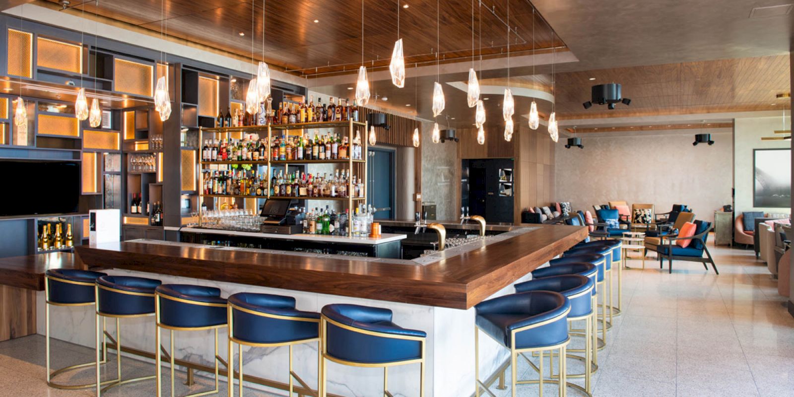 The image shows a modern bar with a wooden countertop, blue chairs, hanging lights, and a well-stocked shelf of bottles in the background.
