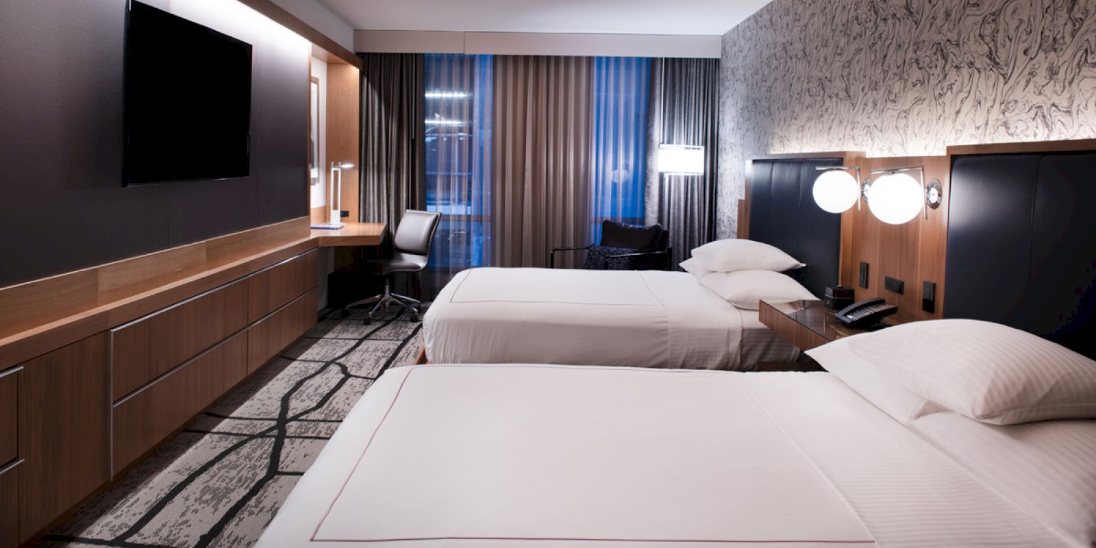 A modern hotel room features two neatly made beds, a desk with a chair, a flat-screen TV on the wall, and stylish decor with a patterned accent wall.