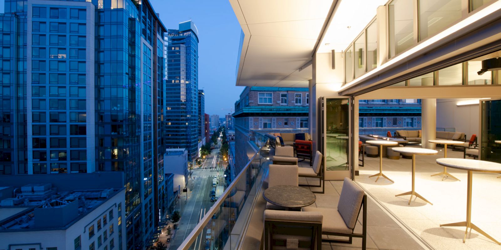 A rooftop terrace with tables and chairs overlooks a cityscape of modern buildings at twilight, featuring a mix of indoor and outdoor lighting.
