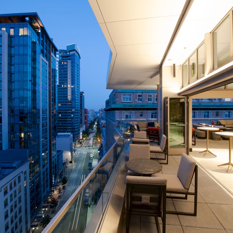 The image shows a modern outdoor patio with seating overlooking a cityscape at dusk, featuring tall glass buildings and a well-lit street.