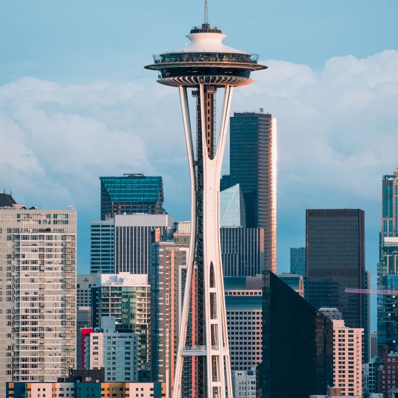 The image shows the Space Needle in Seattle, surrounded by various high-rise buildings against a backdrop of a partially cloudy sky.