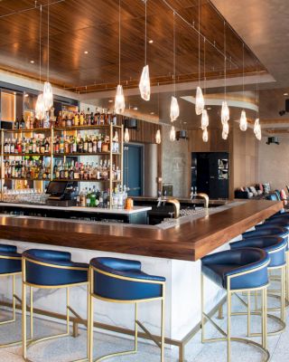The image shows a modern bar with a central island, blue and gold bar stools, shelves of liquor bottles, and hanging pendant lights.