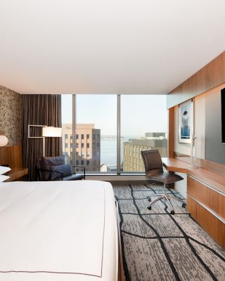 A modern hotel room with a large bed, wooden desk, and flat-screen TV, featuring a city view through large windows and stylish decor.