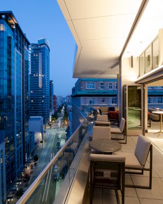 A modern cityscape at dusk, viewed from a balcony with tables and chairs. Tall buildings line the street, windows reflecting the evening sky.