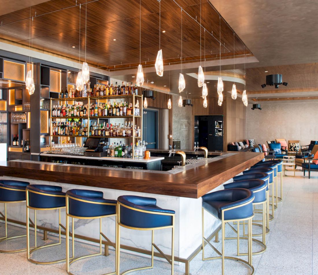 A wide view of a bar with a wooden top, blue stools, and gold accents, with shelves of bottles and glasses in the background.