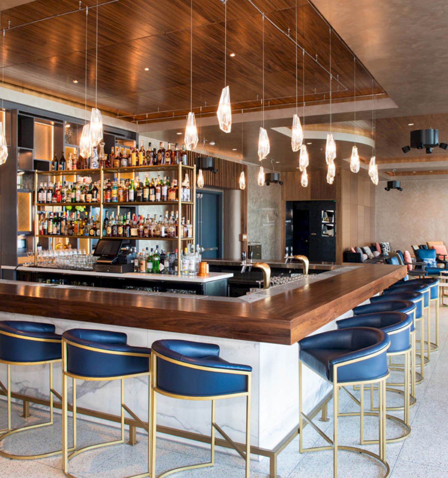 A modern bar with a wooden counter, high stools, pendant lights, and a fully stocked liquor shelf stands in an upscale, spacious interior.