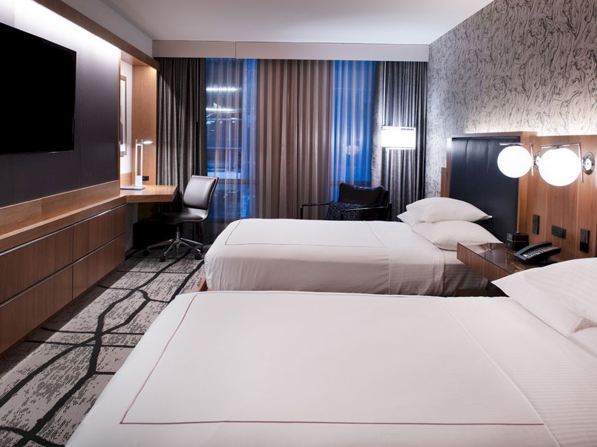 This image shows a modern hotel room with two beds, a flat-screen TV on the wall, a desk with a chair, and lamps beside the beds.