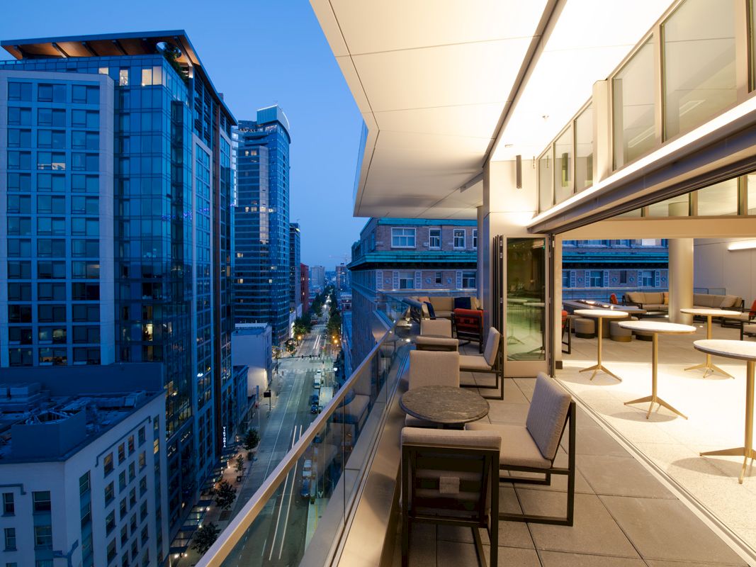 A modern balcony with seating overlooks a city street and tall buildings, illuminated at dusk with a serene urban skyline.