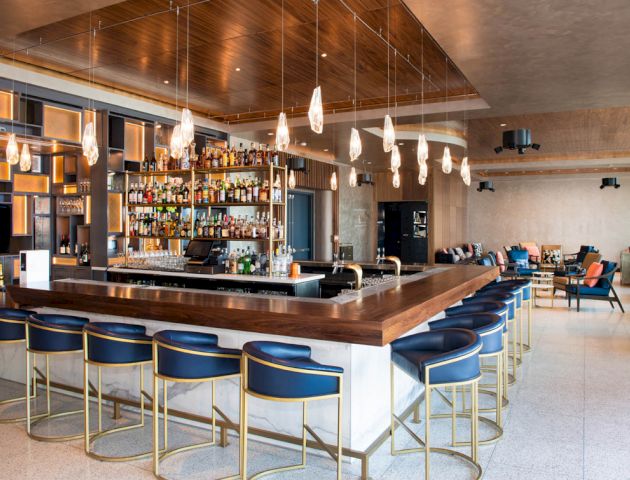 A modern bar with blue seating, a variety of liquor bottles on shelves, pendant lights, and a spacious, sleek design for patrons to enjoy their drinks.