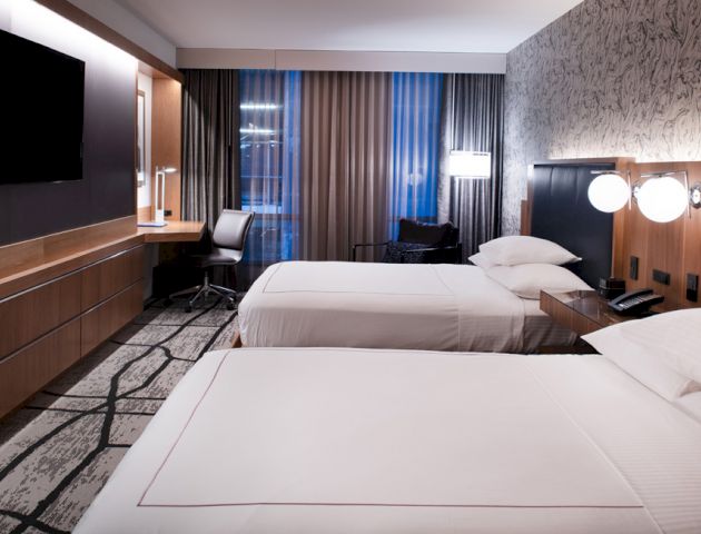 A modern hotel room with two beds, a wall-mounted TV, a desk, and large windows with curtains. The room has contemporary decor and lighting.