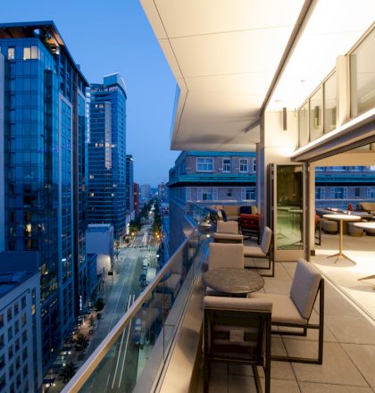 An outdoor balcony with seating and tables, overlooking a cityscape with high-rise buildings at dusk. The area is illuminated by lights.