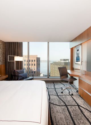 This image shows a modern hotel room featuring a large bed, a desk with a chair, a wall-mounted TV, and a window with a city view.