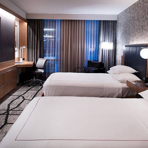 The image shows a modern hotel room with two neatly made beds, a desk area with a chair, a flat-screen TV, and a stylish decor theme.